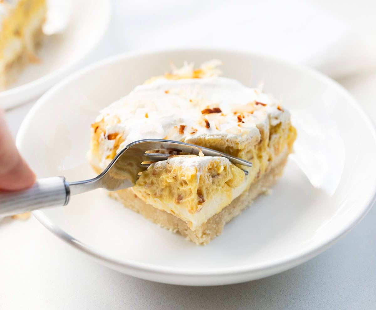 Creamy Coconut Cream Pie Bars with a buttery baked crust, smooth cream cheese layer, and fluffy coconut pudding topped with whipped cream and toasted coconut. Perfect, easy dessert for any occasion! Fork taking a bite of Coconut Cream Pie Bar on a white plate on e white table.
