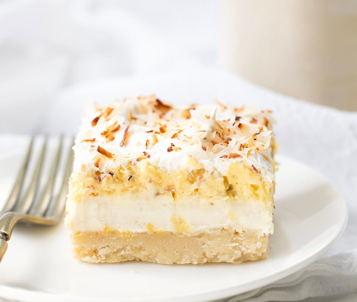 Creamy Coconut Cream Pie Bars with a buttery baked crust, smooth cream cheese layer, and fluffy coconut pudding topped with whipped cream and toasted coconut. Perfect, easy dessert for any occasion! Coconut Cream Pie Bar on a white plate with a fork.