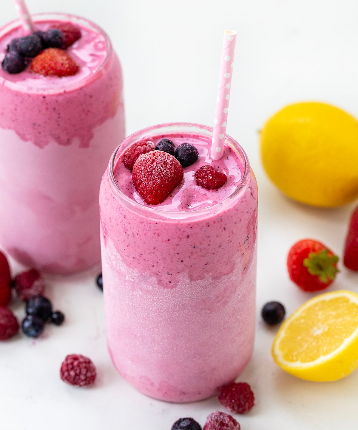 Glasses of Frosted Mixed Berry Lemonade on a white table with fresh and frozen fruit. 