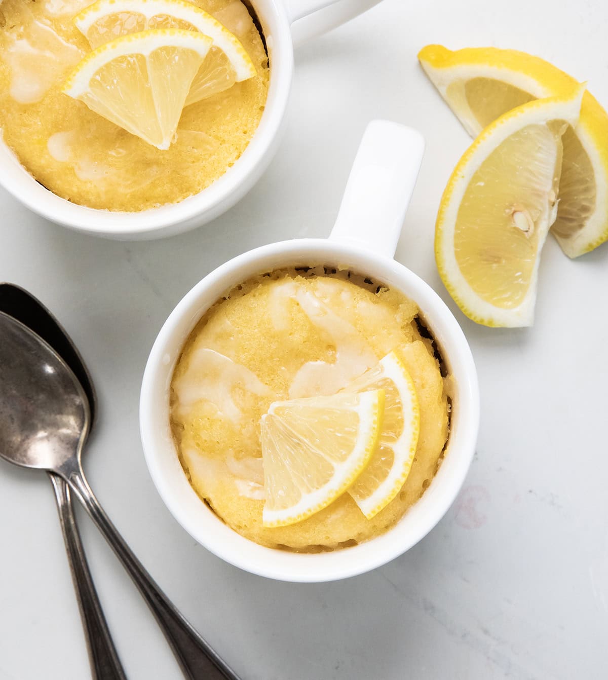 This Lemon Mug Cake is super easy and full of fresh, citrusy flavor. Itโs a quick mug cake recipe you can make in the microwave, perfect when you want a cozy single-serve dessert fast! Soft, sweet, and lemony, this lemon cake totally hits the spot! Lemon Mug Cakes on a white table from overhead.