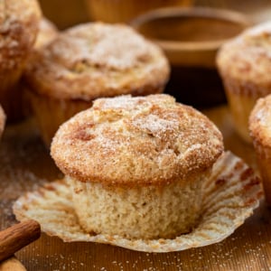Snickerdoodle Muffin on its wrapper on a wooden table surrounded by more muffins and cinnamon sticks.