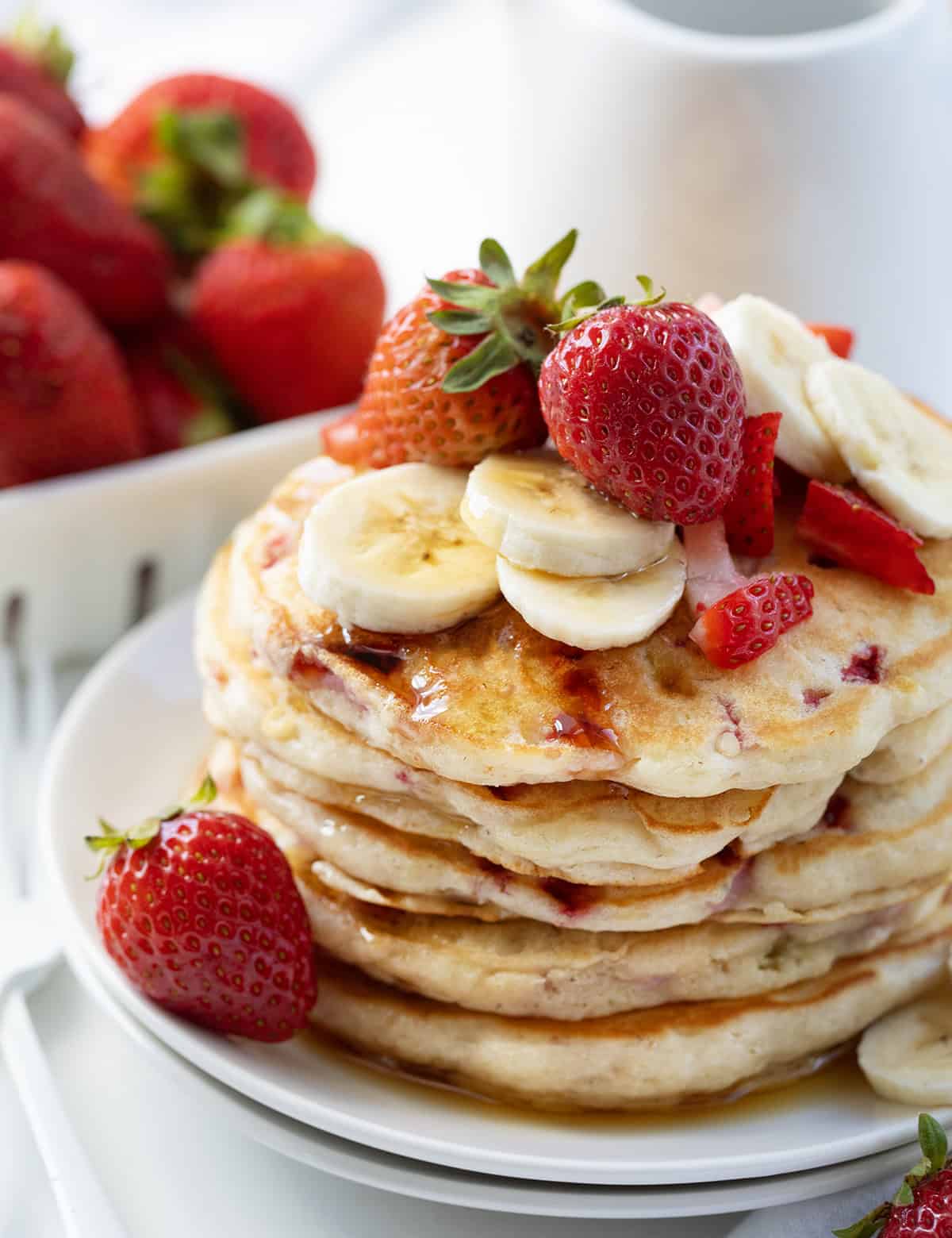 These Strawberry Banana Pancakes are a fruit-filled twist on my classic buttermilk pancakes. Fresh strawberries and mashed bananas are folded right into the batter, making every bite tender, light, and full of flavor! Close up of the top of a stack of Strawberry Banana Pancakes with fresh strawberries and banana slices.
