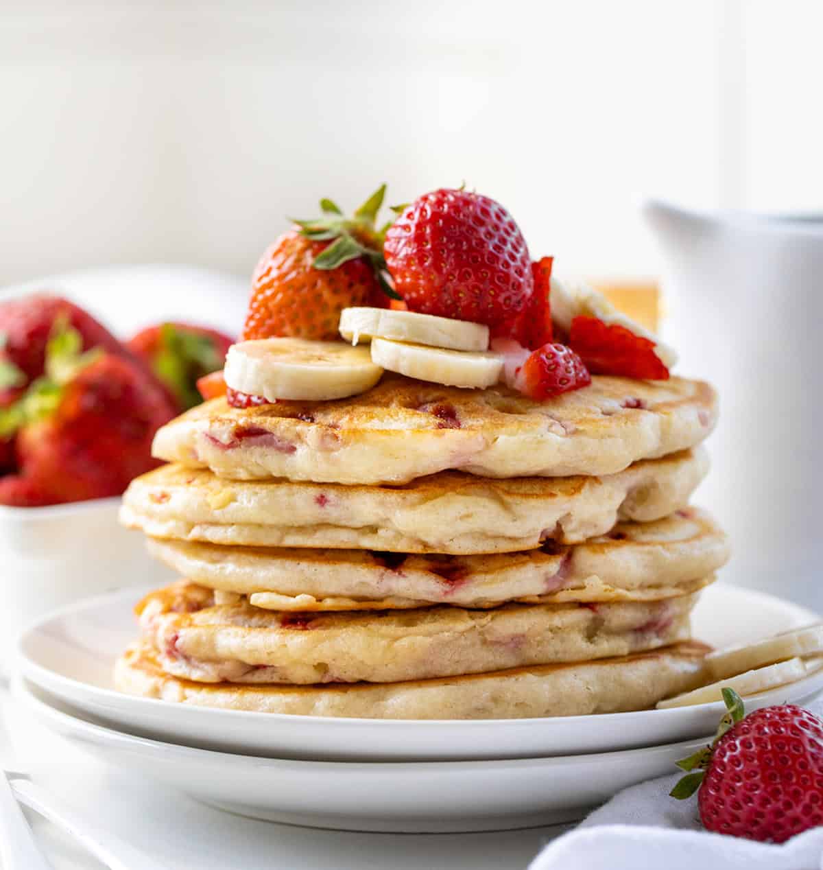 These Strawberry Banana Pancakes are a fruit-filled twist on my classic buttermilk pancakes. Fresh strawberries and mashed bananas are folded right into the batter, making every bite tender, light, and full of flavor! Stack of Strawberry Banana Pancakes on white plates on a white table.