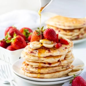 Pouring syrup over Strawberry Banana Pancakes on a white table.