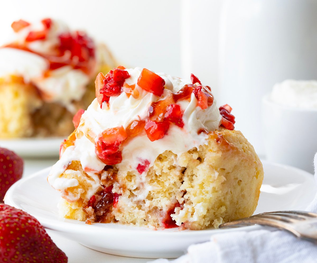 Fluffy biscuit-style cinnamon rolls filled with strawberries and jam, topped with whipped cream and strawberry sauce. These Strawberry Shortcake Cinnamon Rolls give you all the flavors of a classic shortcake in an easy no-rise breakfast or dessert treat! Strawberry Shortcake Cinnamon Roll that has been cut in half on a white plate.