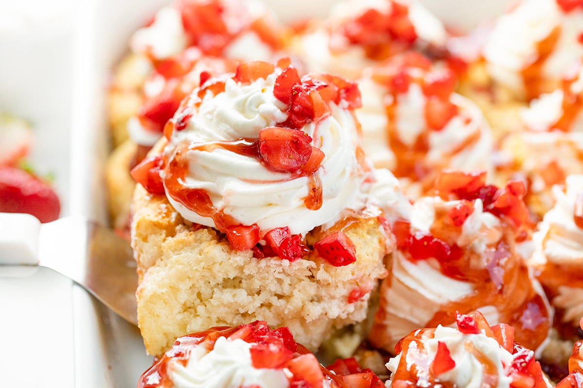 Fluffy biscuit-style cinnamon rolls filled with strawberries and jam, topped with whipped cream and strawberry sauce. These Strawberry Shortcake Cinnamon Rolls give you all the flavors of a classic shortcake in an easy no-rise breakfast or dessert treat! Spatula picking up a Strawberry Shortcake Cinnamon Roll from the pan.