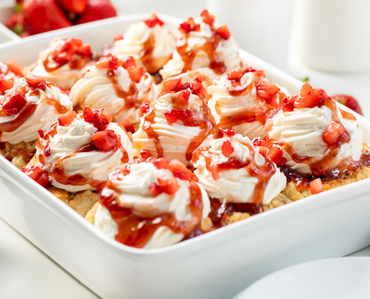 Fluffy biscuit-style cinnamon rolls filled with strawberries and jam, topped with whipped cream and strawberry sauce. These Strawberry Shortcake Cinnamon Rolls give you all the flavors of a classic shortcake in an easy no-rise breakfast or dessert treat! White pan filled with Strawberry Shortcake Cinnamon Rolls on a white counter.