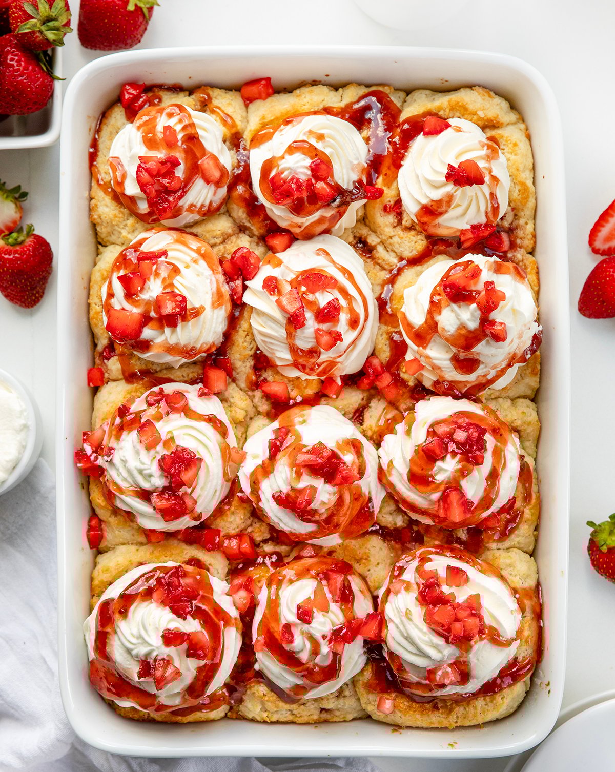 Fluffy biscuit-style cinnamon rolls filled with strawberries and jam, topped with whipped cream and strawberry sauce. These Strawberry Shortcake Cinnamon Rolls give you all the flavors of a classic shortcake in an easy no-rise breakfast or dessert treat! White pan with Strawberry Shortcake Cinnamon Rolls on a white counter from overhead.