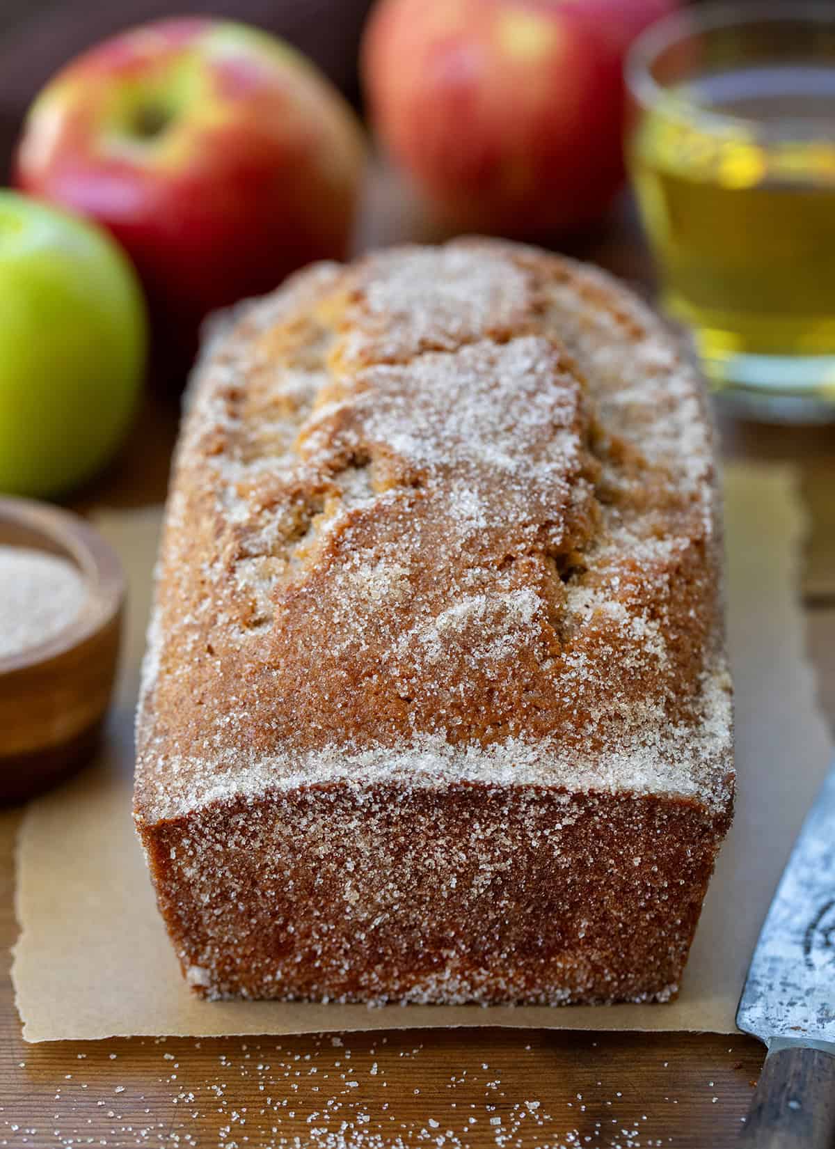 This Apple Cider Donut Bread is tender, cozy, and full of apple cider flavor, with warm apple pie spice in every bite. After baking, I pour apple cider over the warm loaf and sprinkle it with a cinnamon-sugar topping for that perfect “donut” finish... slightly crisp on the outside, soft and moist inside! Loaf of Apple Cider Donut Bread on a wooden table with fresh apples and apple cider.