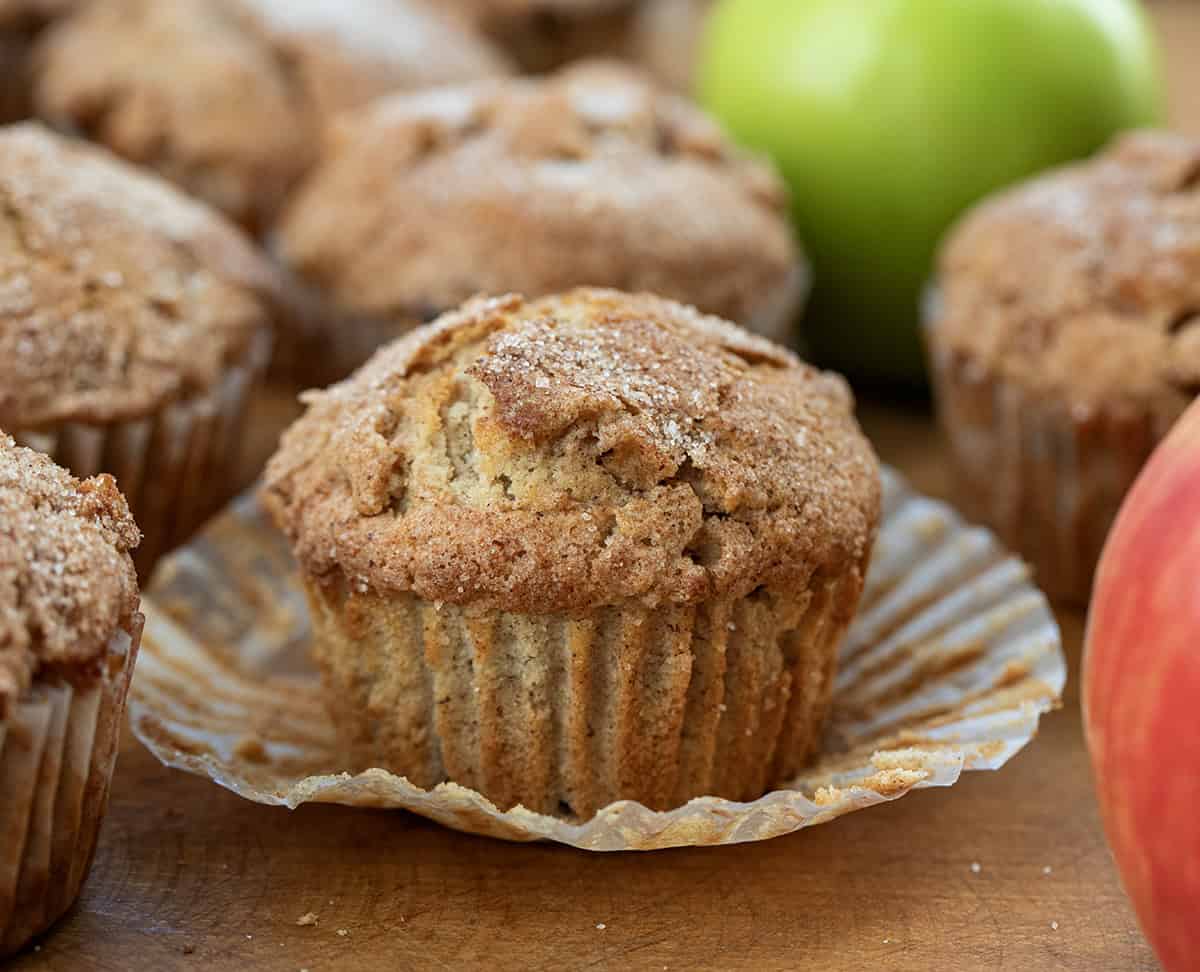 These Apple Snickerdoodle Muffins are soft, fluffy, and full of cozy fall flavor with apples, warm spice, and a buttery cinnamon-sugar topping. Perfect for breakfast, brunch, or snacking! Apple Snickerdoodle Muffins on a wooden table with center muffin unwrapped.