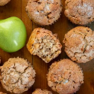 Apple Snickerdoodle Muffins on a wooden table with the center muffin halved and showing inside texture.