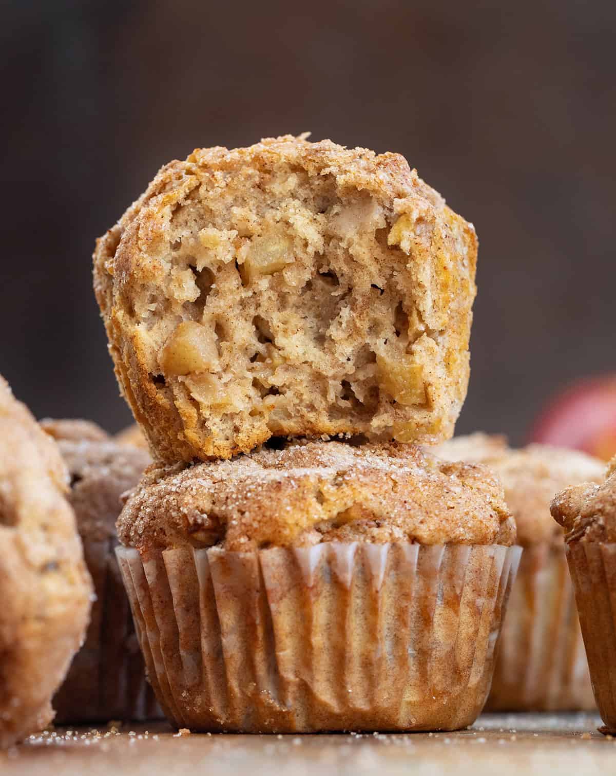 These Apple Snickerdoodle Muffins are soft, fluffy, and full of cozy fall flavor with apples, warm spice, and a buttery cinnamon-sugar topping. Perfect for breakfast, brunch, or snacking! Stack of Apple Snickerdoodle Muffins on a wooden table with an apple in the background.