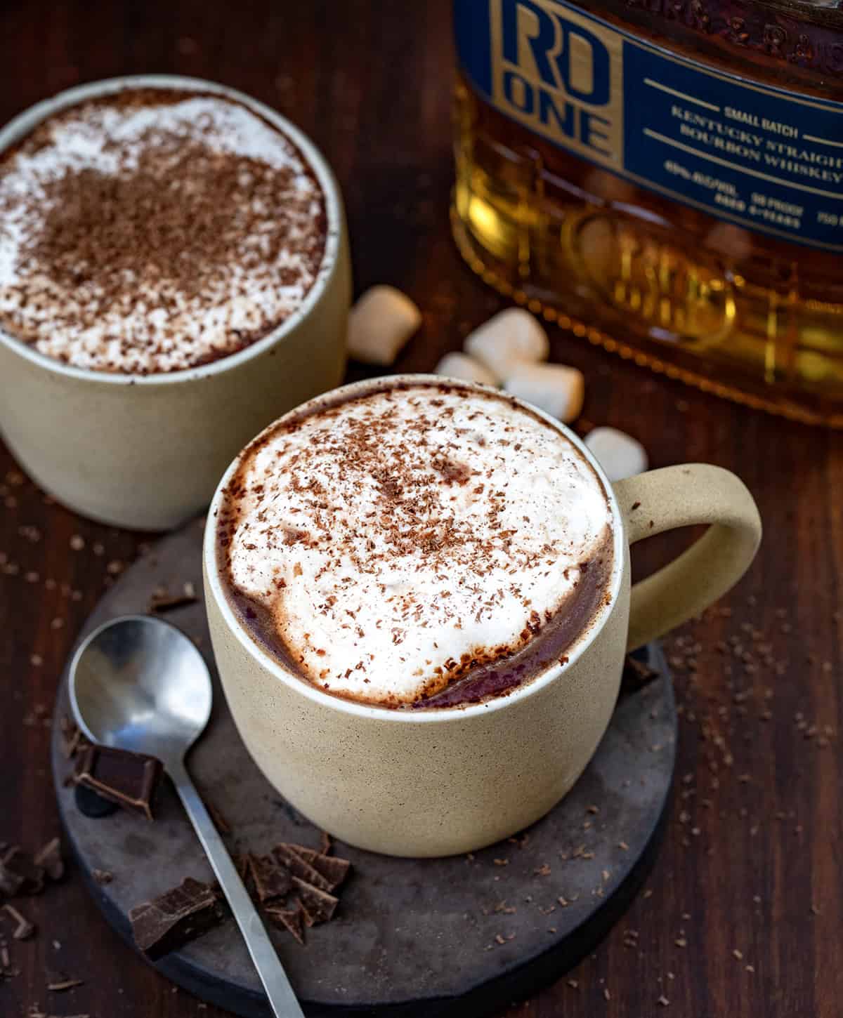 Glasses of Bad Santa Hot Chocolate on a wooden table with chopped chocolate, a mini spoon, and a bottle of bourbon.