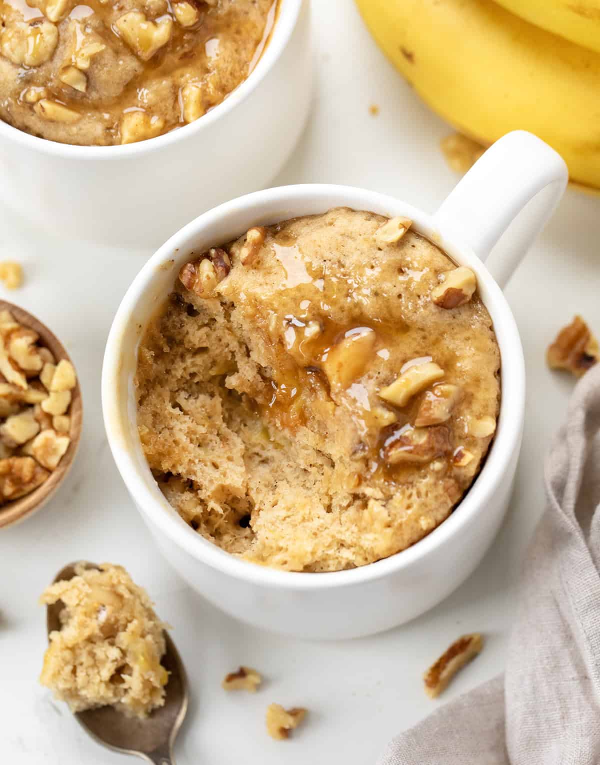 Close up of a white mug with Banana Nut Mug Cake on a cutting board with a bite taken out of it and fresh bananas.