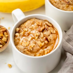 Close up of a white mug with Banana Nut Mug Cake on a cutting board with fresh bananas.