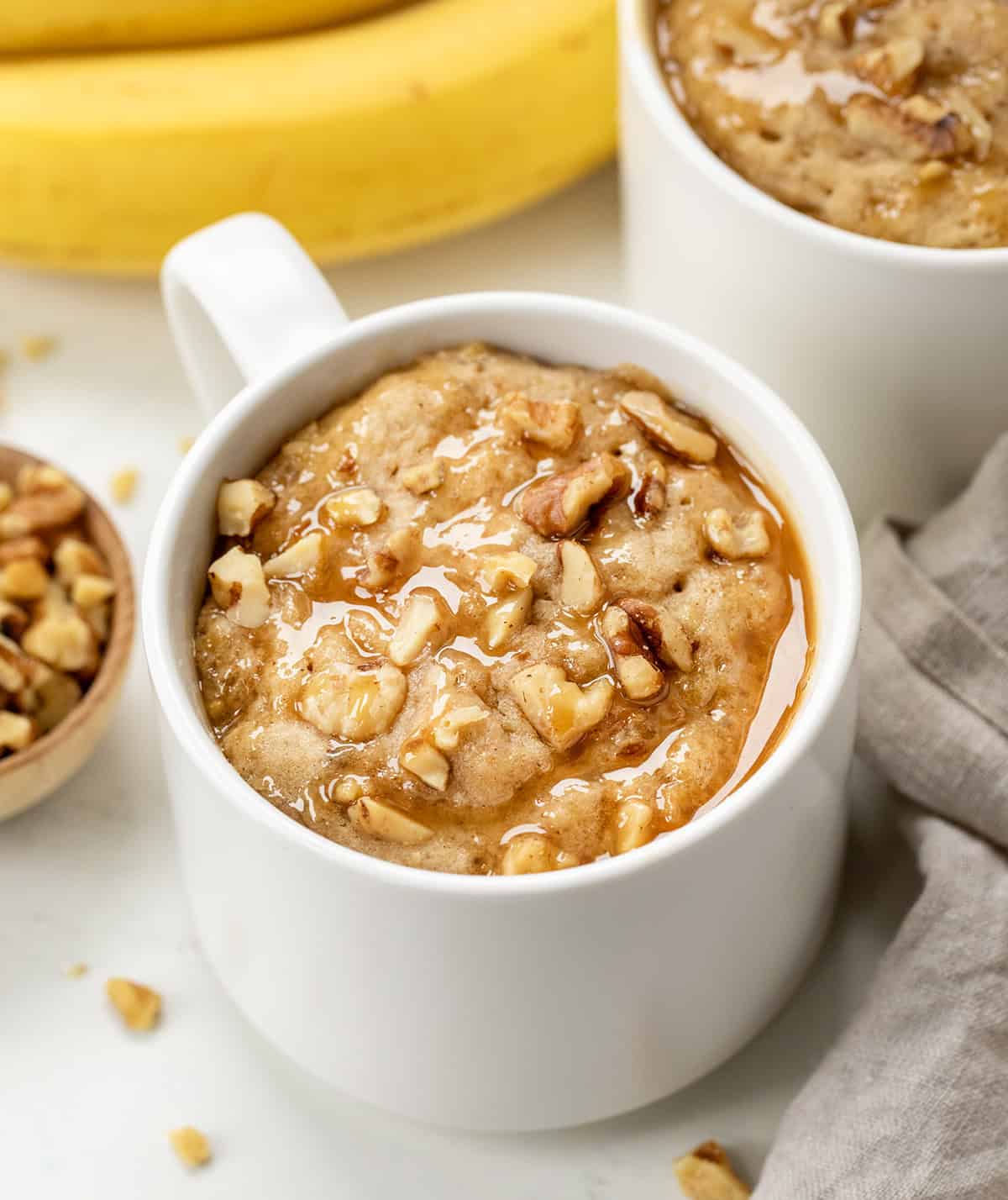 Close up of a white mug with Banana Nut Mug Cake on a cutting board with fresh bananas.