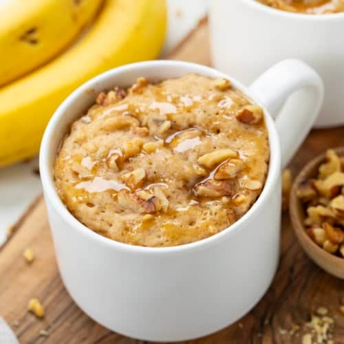 Close up of a white mug with Banana Nut Mug Cake on a cutting board with fresh bananas.