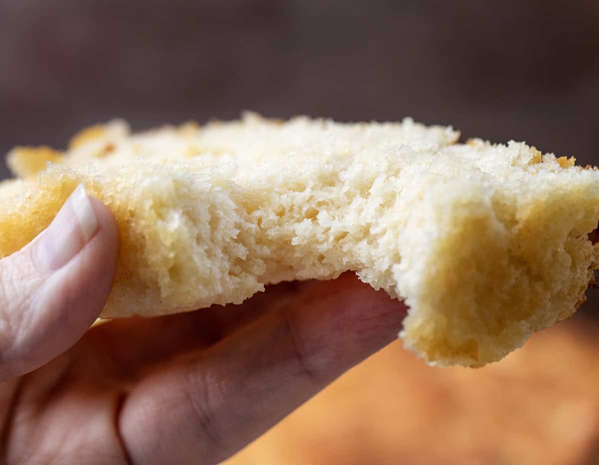This Butter Swim Bread is golden, buttery, and irresistibly tender because it bakes right in a pool of melted butter. The butter gives the loaf crisp edges, a rich flavor, and a soft, biscuit-like crumb that melts in your mouth! Hand holding a piece of Butter Swim Bread with a bite taken out showing tender crumb.