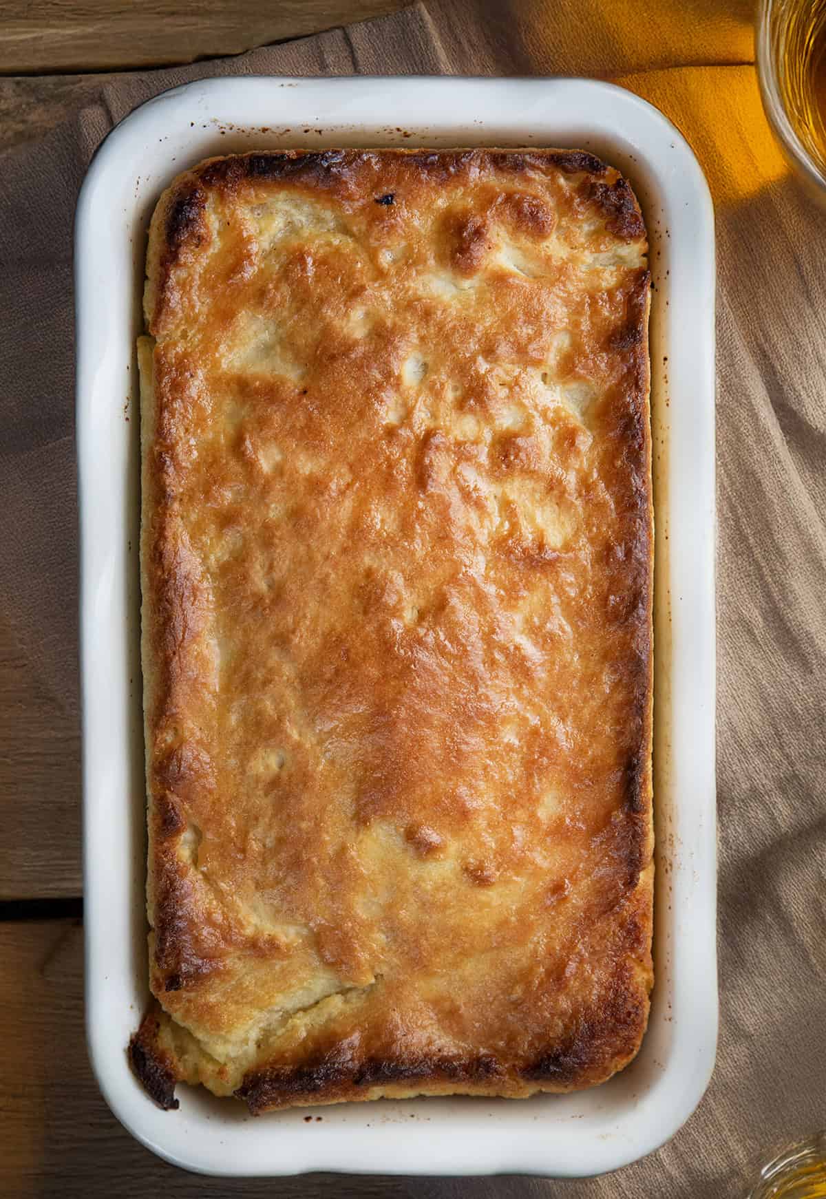 This Butter Swim Bread is golden, buttery, and irresistibly tender because it bakes right in a pool of melted butter. The butter gives the loaf crisp edges, a rich flavor, and a soft, biscuit-like crumb that melts in your mouth! Loaf of Butter Swim Bread in a white pan on a wooden table from overhead.