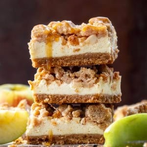 Stack of Caramel Apple Cheesecake Bars on a wooden table with fresh apples.
