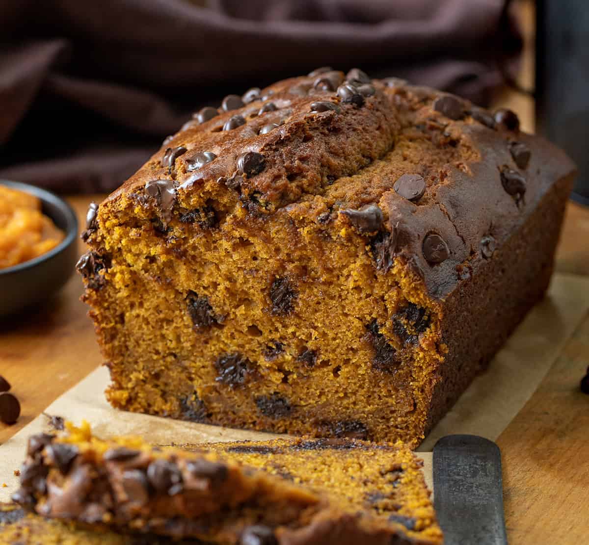 This Chocolate Chip Pumpkin Bread is soft, tender, and loaded with melty chocolate chips inside and on top. Sweet enough to feel like a treat but still full of that cozy pumpkin flavor we all love, itโs the perfect quick bread for fall baking! Loaf of Chocolate Chip Pumpkin Bread that has been cut into on a wooden table.
