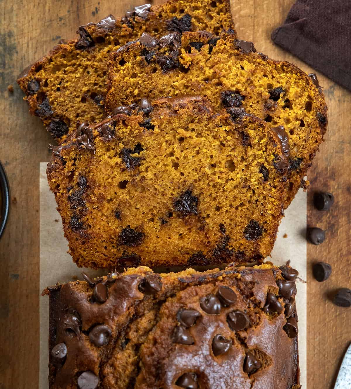 This Chocolate Chip Pumpkin Bread is soft, tender, and loaded with melty chocolate chips inside and on top. Sweet enough to feel like a treat but still full of that cozy pumpkin flavor we all love, itโs the perfect quick bread for fall baking! Loaf of Chocolate Chip Pumpkin Bread with slices cut and fallen, laying flat on a wooden table.