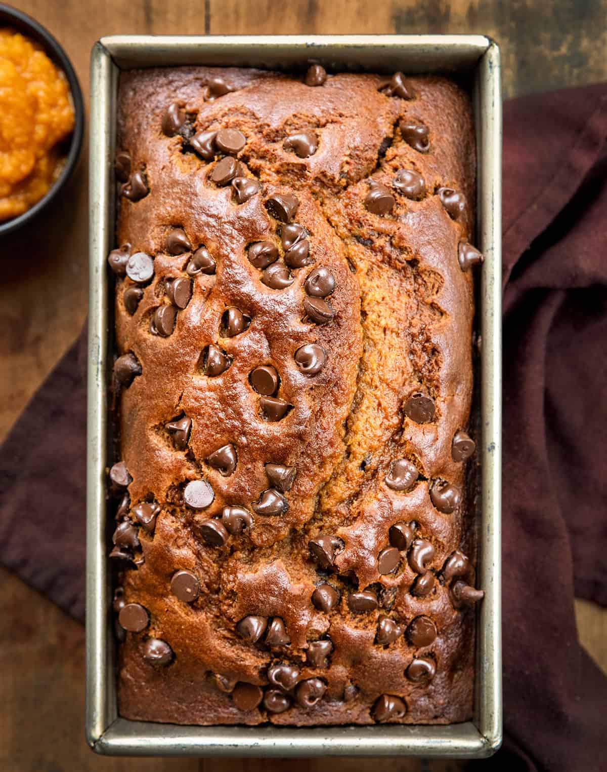 This Chocolate Chip Pumpkin Bread is soft, tender, and loaded with melty chocolate chips inside and on top. Sweet enough to feel like a treat but still full of that cozy pumpkin flavor we all love, itโs the perfect quick bread for fall baking! Loaf of Chocolate Chip Pumpkin Bread in the pan on a wooden table from overhead.