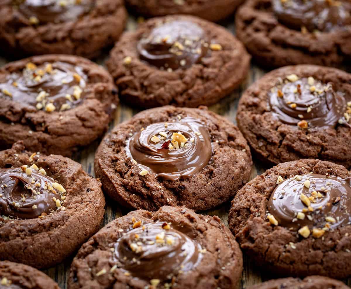 These Chocolate Hazelnut Thumbprint Cookies are rich, fudgy, and filled with creamy chocolate-hazelnut spread (such as Nutella!). The cookies are soft in the center with slightly crisp edges, and the gooey filling makes them totally irresistible. Chocolate Hazelnut Thumbprint Cookies close together with chopped hazelnut pieces sprinkled over the Nutella.