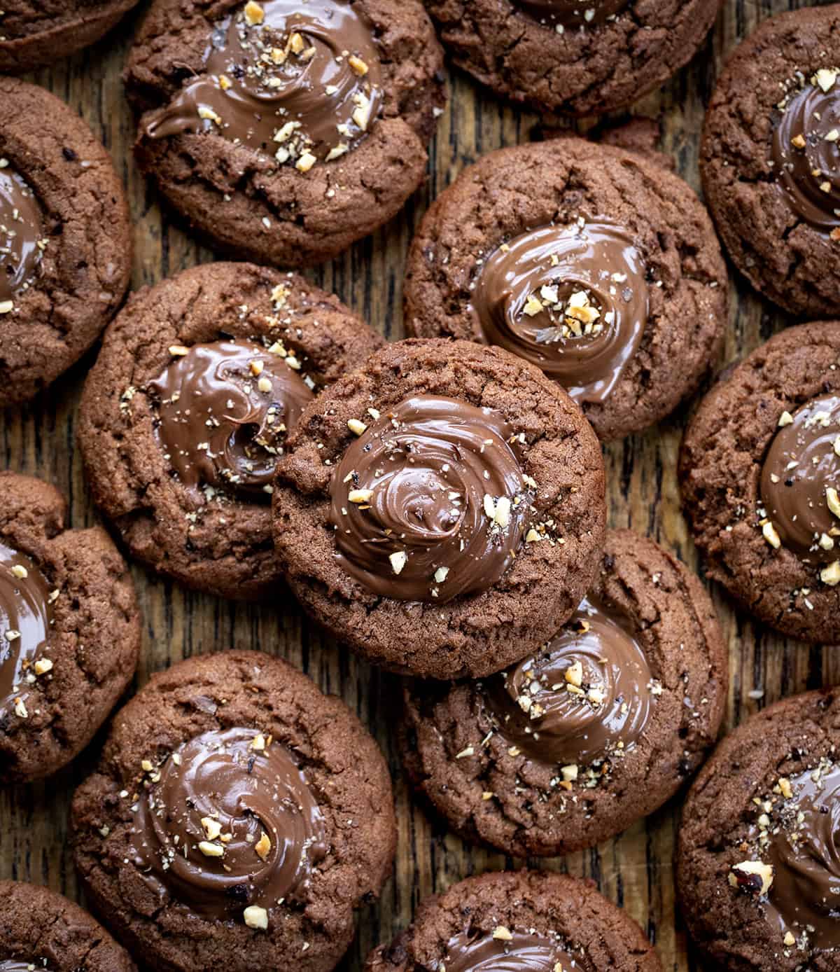 These Chocolate Hazelnut Thumbprint Cookies are rich, fudgy, and filled with creamy chocolate-hazelnut spread (such as Nutella!). The cookies are soft in the center with slightly crisp edges, and the gooey filling makes them totally irresistible. Chocolate Hazelnut Thumbprint Cookies on a wooden table with a couple of the cookies stacked.