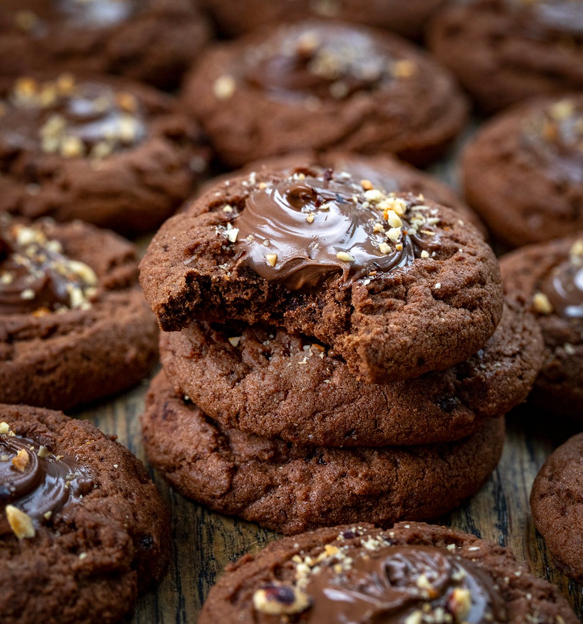 These Chocolate Hazelnut Thumbprint Cookies are rich, fudgy, and filled with creamy chocolate-hazelnut spread (such as Nutella!). The cookies are soft in the center with slightly crisp edges, and the gooey filling makes them totally irresistible. Stack of Chocolate Hazelnut Thumbprint Cookies with the top cookie bit into showing tender texture.