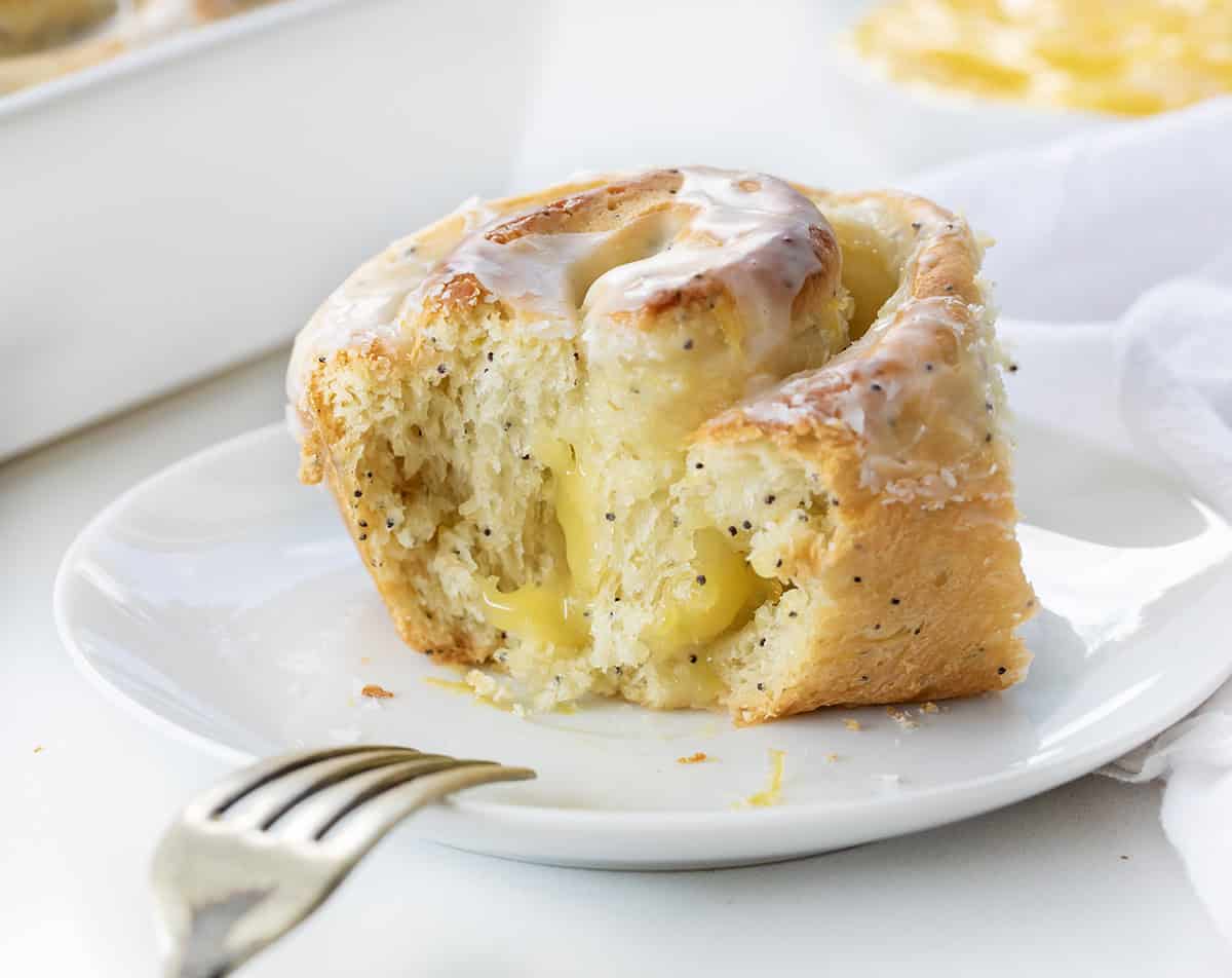 These Lemon Poppy Seed Rolls are soft, fluffy, sweet rolls filled with tangy lemon curd and topped with a light glaze that soaks into every bite! Lemon Poppy Seed Roll on a white plate with a portion of the roll removed showing inside texture.