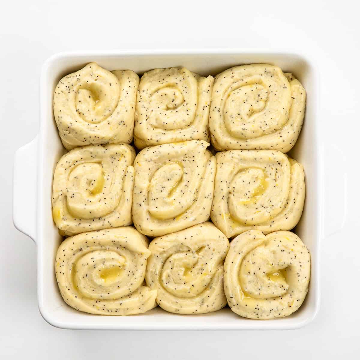 These Lemon Poppy Seed Rolls are soft, fluffy, sweet rolls filled with tangy lemon curd and topped with a light glaze that soaks into every bite! Lemon Poppy Seed Rolls before being baked in a white pan on a white table.