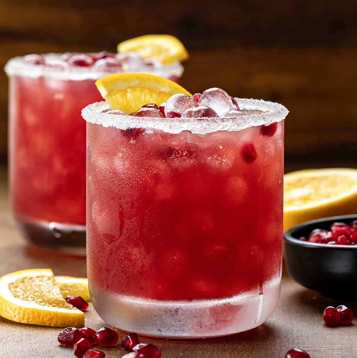 Close up of glasses of Orange Pomegranate Margarita on a wooden table with fresh orange and pomegranate. 