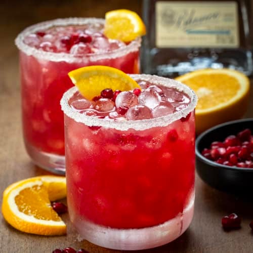 Close up of glasses of Orange Pomegranate Margarita on a wooden table with fresh orange and pomegranate.