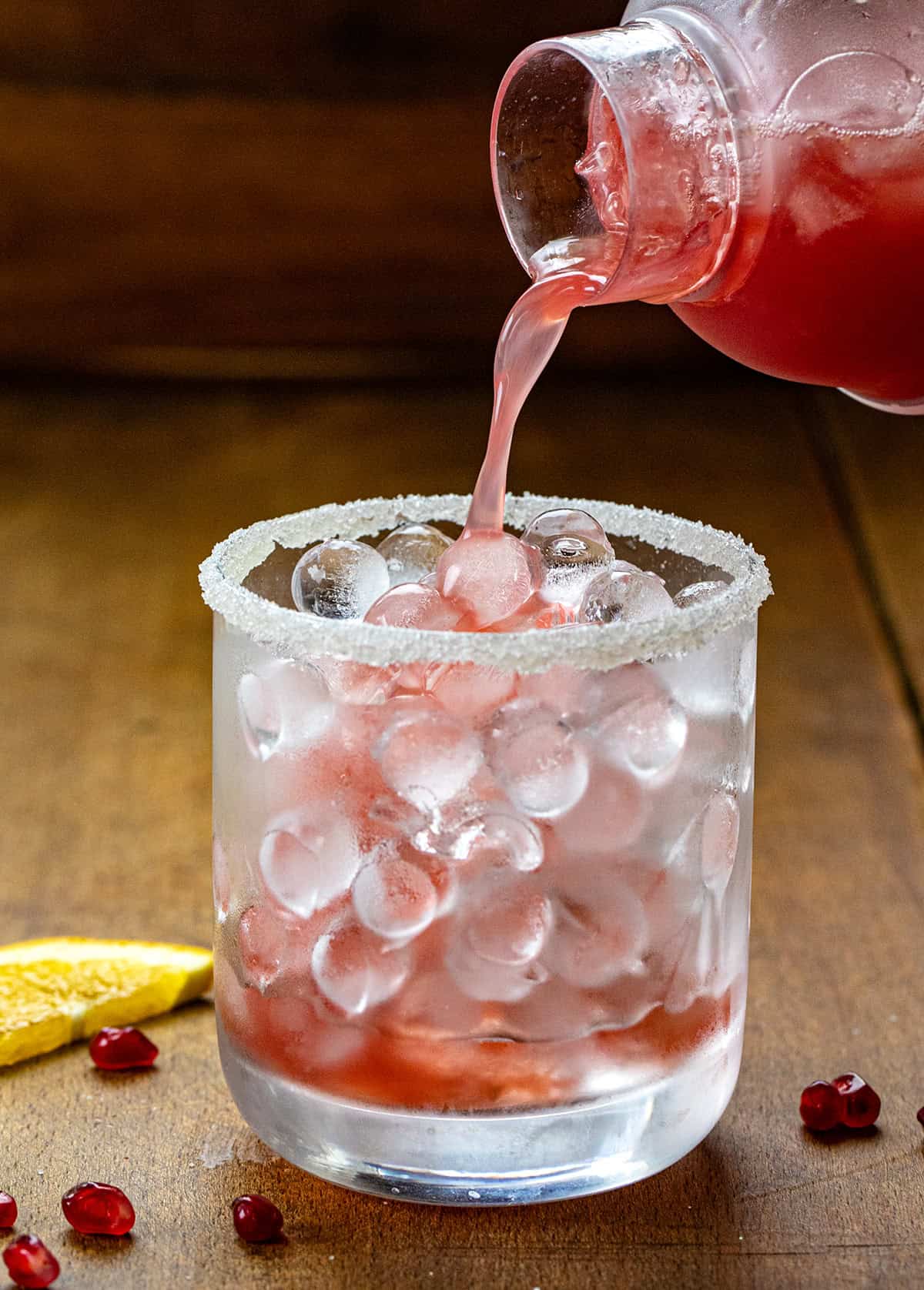 Pourning Orange Pomegranate Margarita into a glass with small round ice cubes on a wooden table.