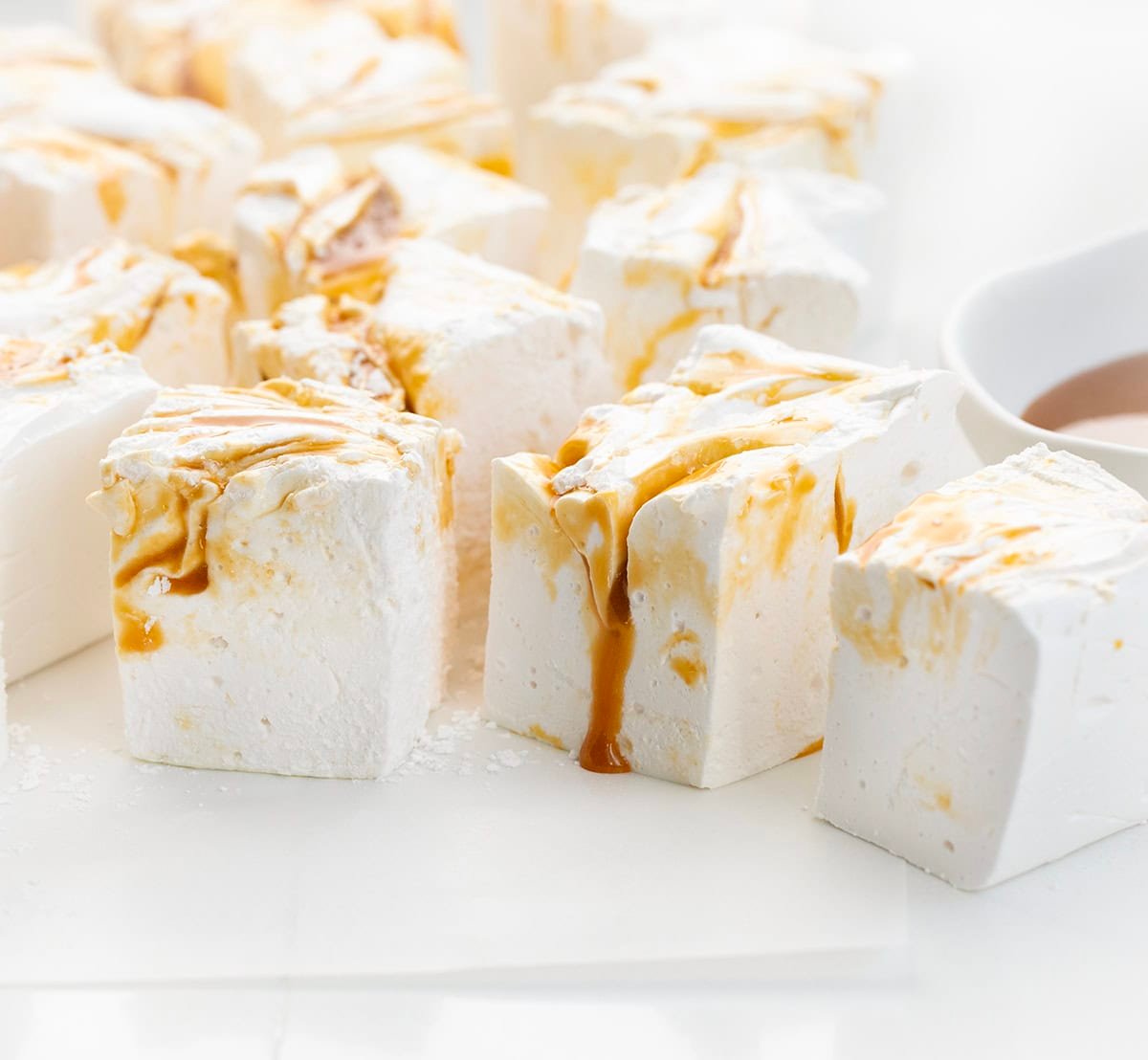 Salted Caramel Swirl Marshmallows cut into squares on a white table with a bowl of caramel sauce.