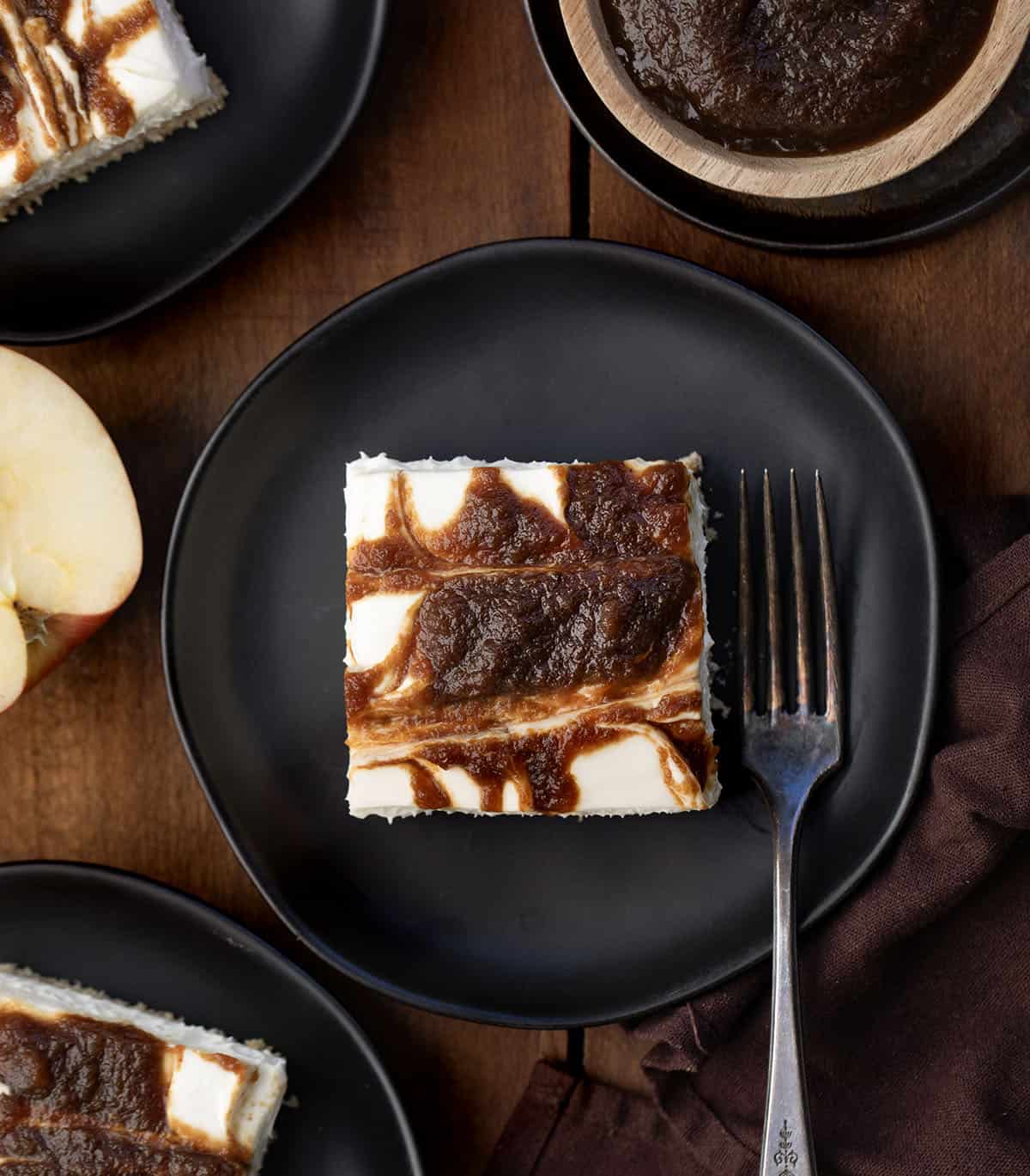 If you love my Apple Butter Cheesecake, you’re going to adore these Apple Butter Cheesecake Bars! With a buttery graham cracker crust, fluffy cream cheese filling, and swirls of sweet apple butter, these bars are perfectly rich, yet so light! Apple Butter Cheesecake Bars cut into squares on black plates on a wooden table from overhead.