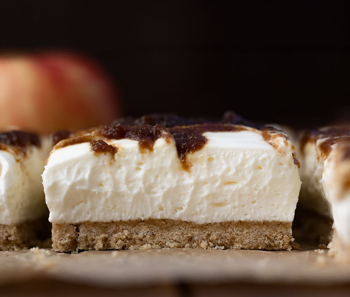 If you love my Apple Butter Cheesecake, you’re going to adore these Apple Butter Cheesecake Bars! With a buttery graham cracker crust, fluffy cream cheese filling, and swirls of sweet apple butter, these bars are perfectly rich, yet so light! Close up of a Apple Butter Cheesecake Bar on a piece of brown parchment paper and an apple in the background.