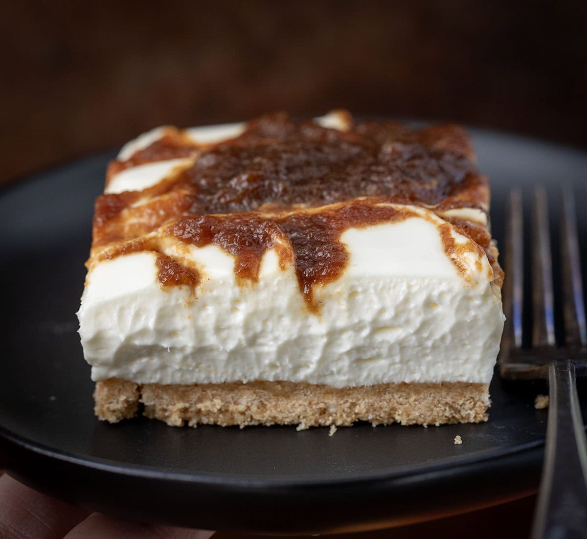If you love my Apple Butter Cheesecake, you’re going to adore these Apple Butter Cheesecake Bars! With a buttery graham cracker crust, fluffy cream cheese filling, and swirls of sweet apple butter, these bars are perfectly rich, yet so light! Apple Butter Cheesecake Bar on a black plate with a fork.