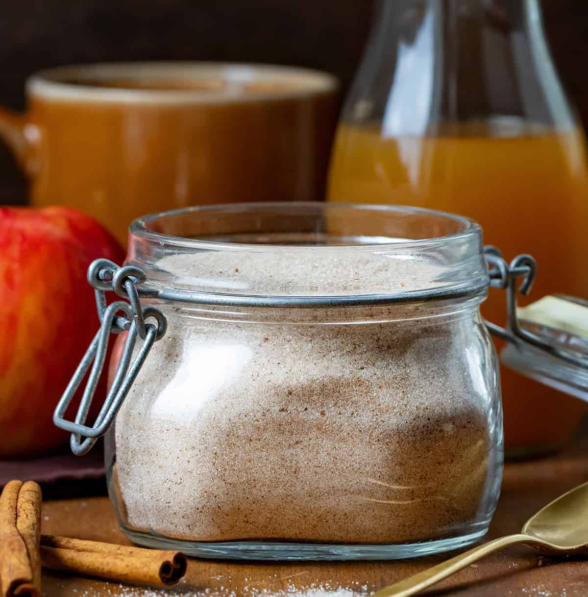 Make your own spiced Apple Cider Mix to have cozy fall flavors ready anytime! This easy blend of sugar and apple pie spice turns plain apple juice into a warm, comforting drink perfect for chilly mornings, holiday gatherings, or just a cozy treat anytime. Jar of Apple Cider Mix on a wooden table with cider, an apple, a spoon, cinnamon sticks, and a mug.