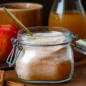 Jar of Apple Cider Mix on a wooden table with cider, an apple, a spoon, cinnamon sticks, and a mug.