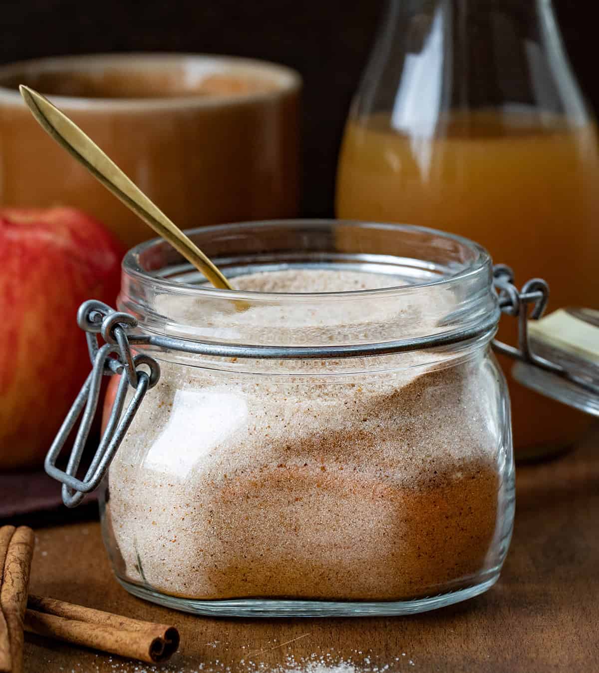 Jar of Apple Cider Mix on a wooden table with cider, an apple, a spoon, cinnamon sticks, and a mug.
