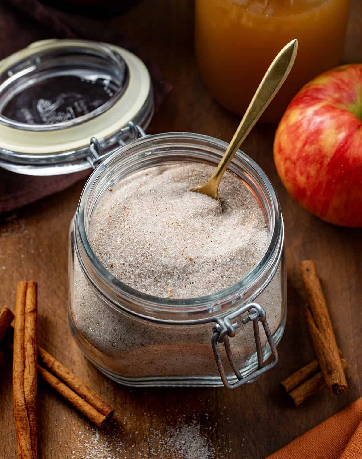 Make your own spiced Apple Cider Mix to have cozy fall flavors ready anytime! This easy blend of sugar and apple pie spice turns plain apple juice into a warm, comforting drink perfect for chilly mornings, holiday gatherings, or just a cozy treat anytime. Jar of Apple Cider Mix on a wooden table with cider, an apple, a spoon, cinnamon sticks, and a mug.