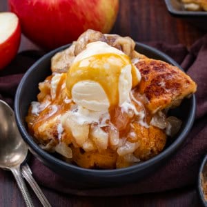 Bowl of Apple Cinnamon Roll Bake on a wooden table with spoons and apples.