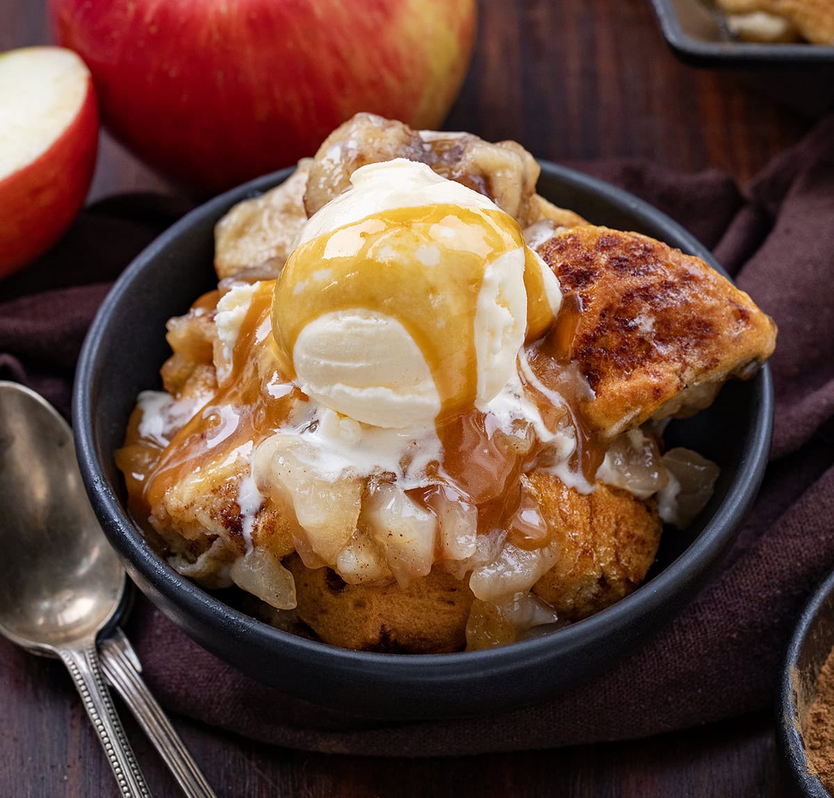 This 2-Ingredient Apple Cinnamon Roll Bake is effortless, cozy, and perfect for a snack, brunch, or dessert! Using store-bought cinnamon rolls and sweet apple pie filling, this bake comes together in just minutes but tastes like a homemade treat. Bowl of Apple Cinnamon Roll Bake on a wooden table with spoons and apples.