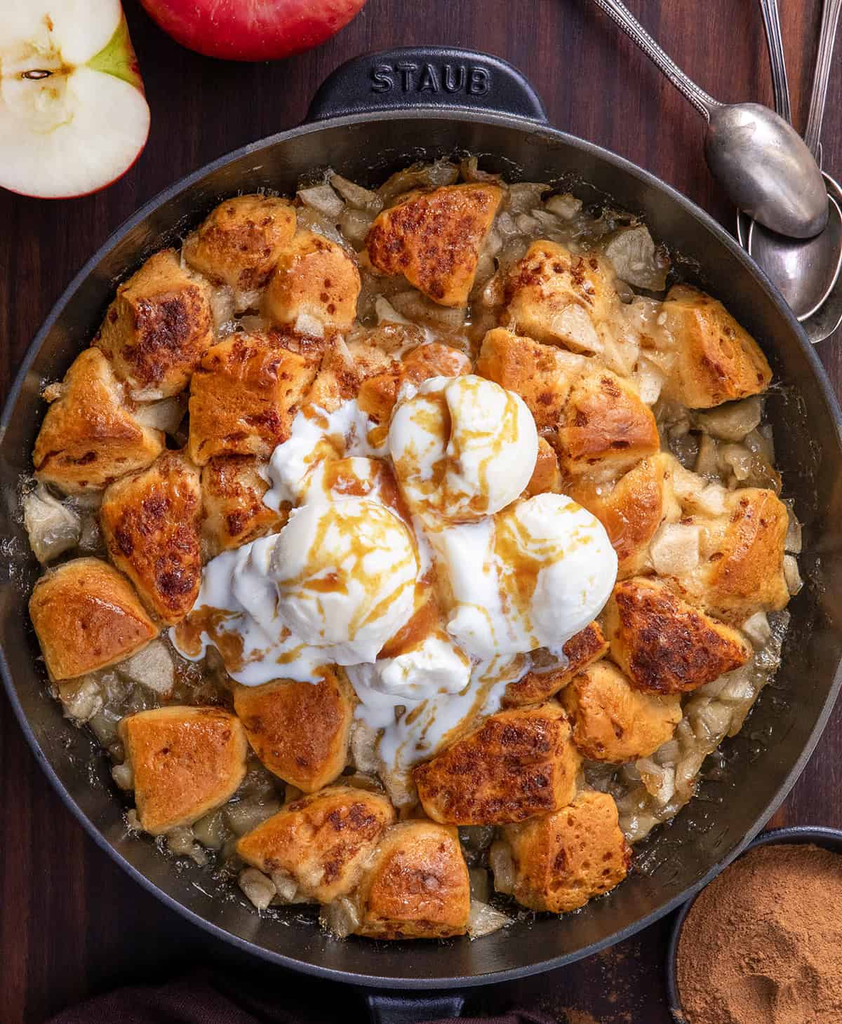 This 2-Ingredient Apple Cinnamon Roll Bake is effortless, cozy, and perfect for a snack, brunch, or dessert! Using store-bought cinnamon rolls and sweet apple pie filling, this bake comes together in just minutes but tastes like a homemade treat. Skillet of Apple Cinnamon Roll Bake with ice cream on top on a wooden table from overhead.