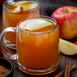 Glass mugs of Maple Bourbon Apple Cider on a wooden table with fresh apples and cinnamon.