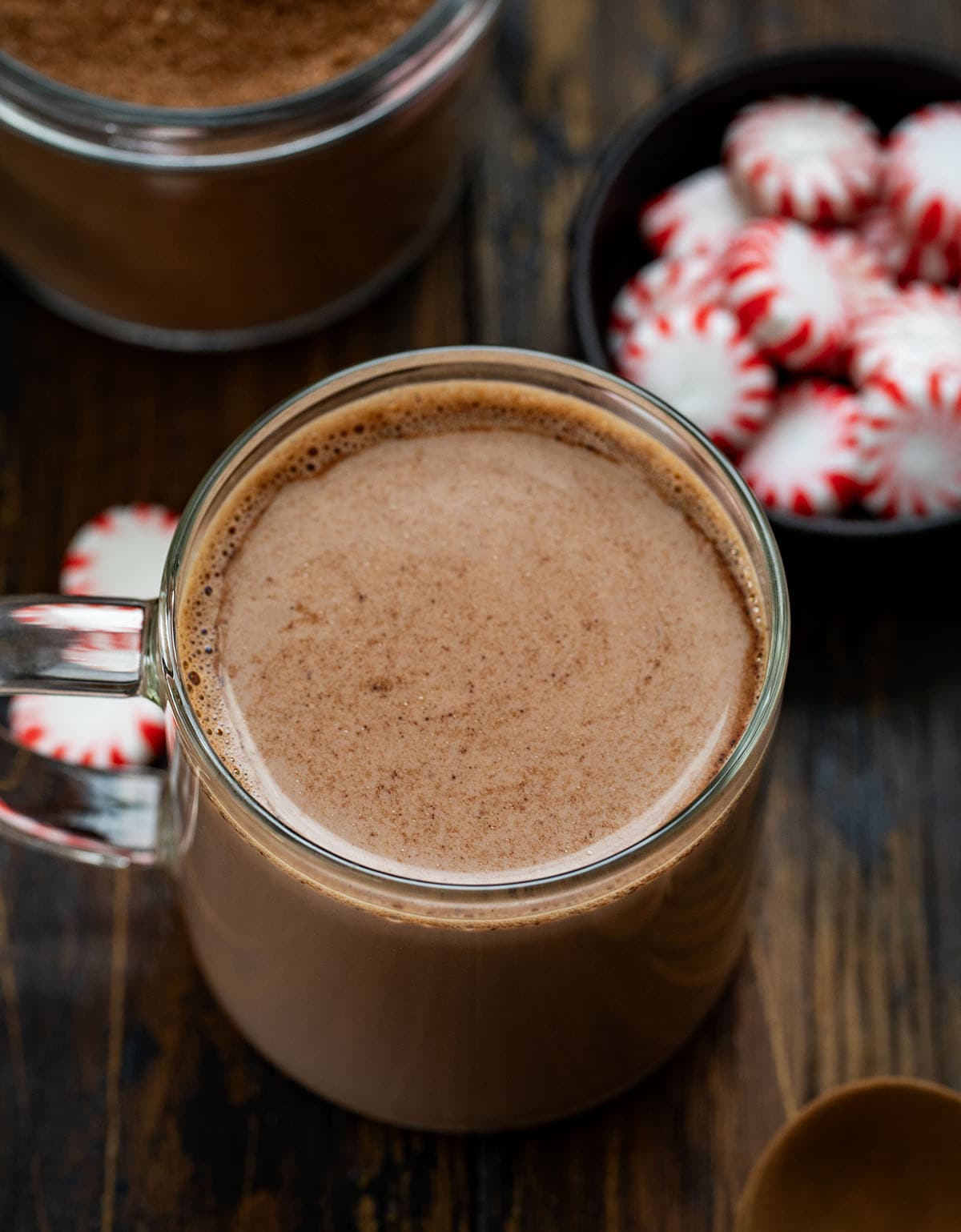 Nothing says cozy winter nights like a mug of rich, creamy hot chocolate with a hint of peppermint. This Peppermint Hot Cocoa Mix is easy to make ahead, customizable, and perfect for gifting during the holidays. Each sip delivers a smooth chocolate flavor with a refreshing minty finish! Glass mug of Peppermint Hot Cocoa on a wooden table with mints and the Peppermint Hot Cocoa Mix behind it.