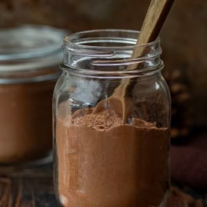 Jar of Peppermint Hot Cocoa Mix on a wooden table with a wooden measuring spoon in the jar.