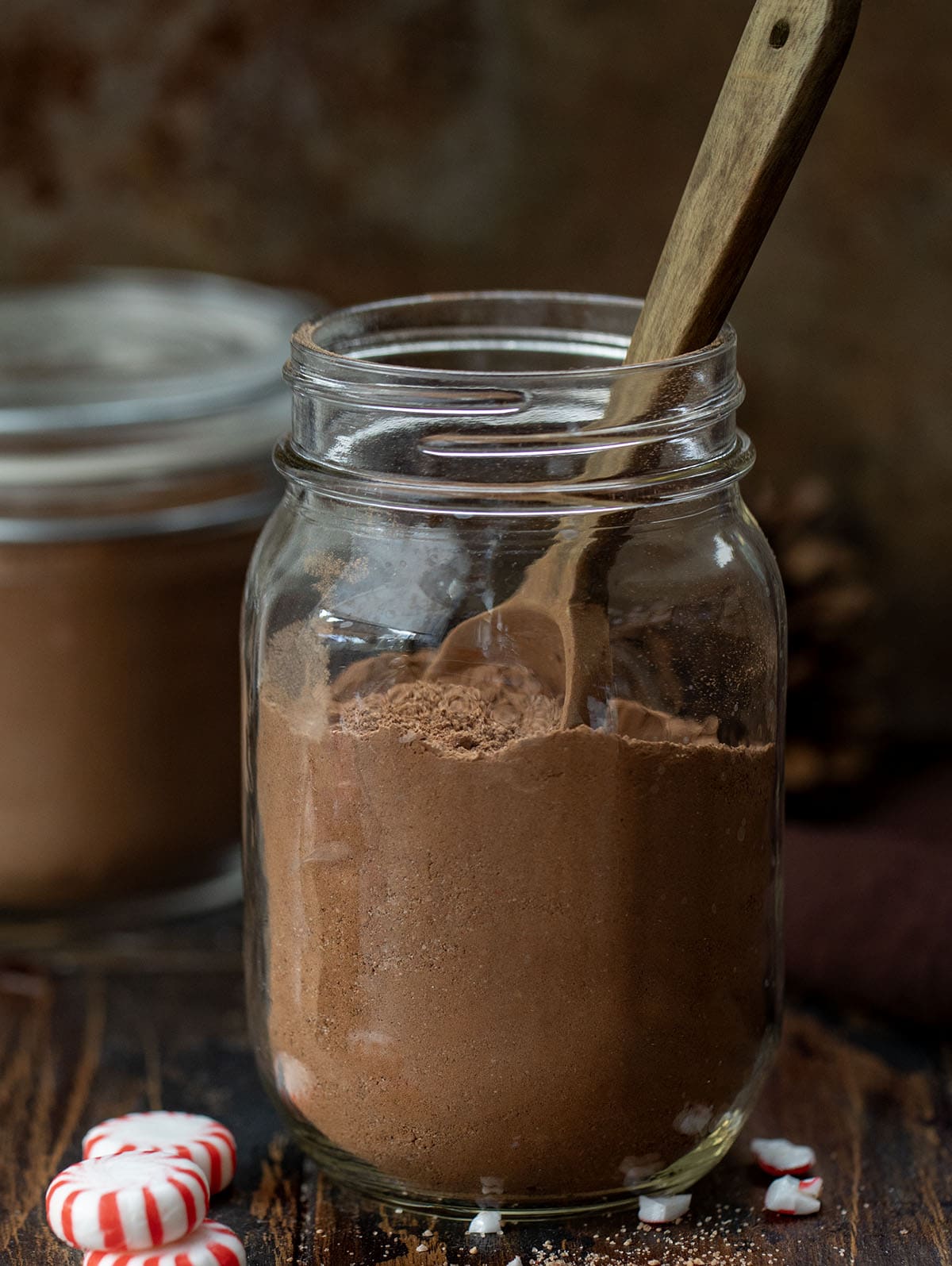Jar of Peppermint Hot Cocoa Mix on a wooden table with a wooden measuring spoon in the jar.
